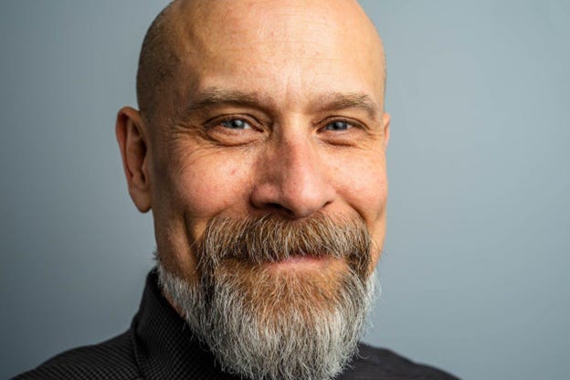 Bald, older man with graying beard against slate background looking at camera