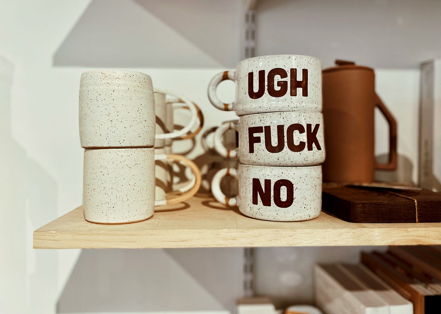 A photo of a wooden shelf against a white background. On the shelf are a stack of white mugs. Three of them have words on them that say: ugh, fuck, no