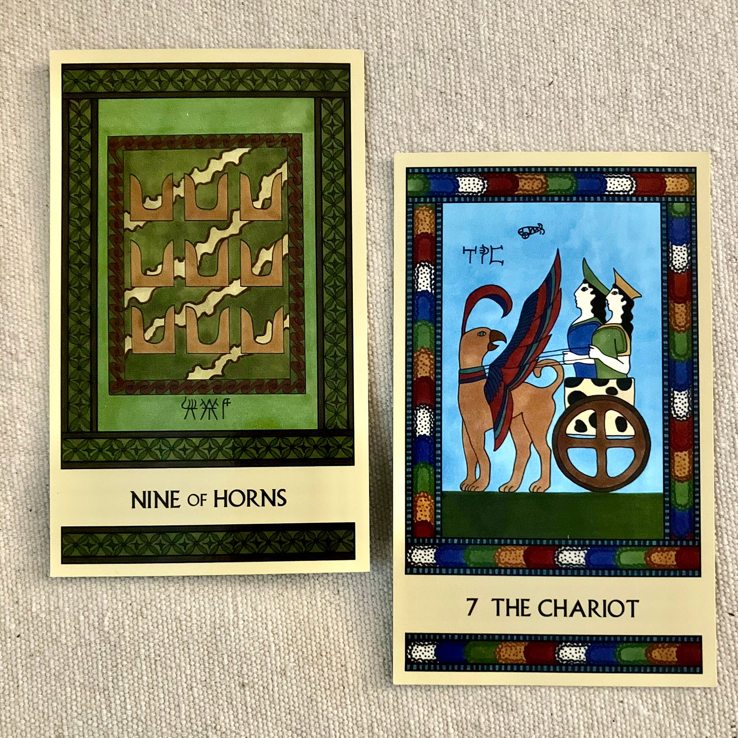 Two Minoan Tarot cards on an off white canvas background. The Nine of Horns is in shades of green and brown. It shows a fancy Minoan fresco with nine pairs of sacred horns painted on it. The Chariot has a multi-colored border. It shows two goddesses riding on a chariot pulled by a griffin.