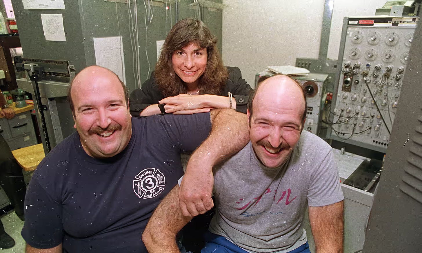 Two identical twins, bald, mustached, smiling. A brunette woman behind them. Set in a fire station.