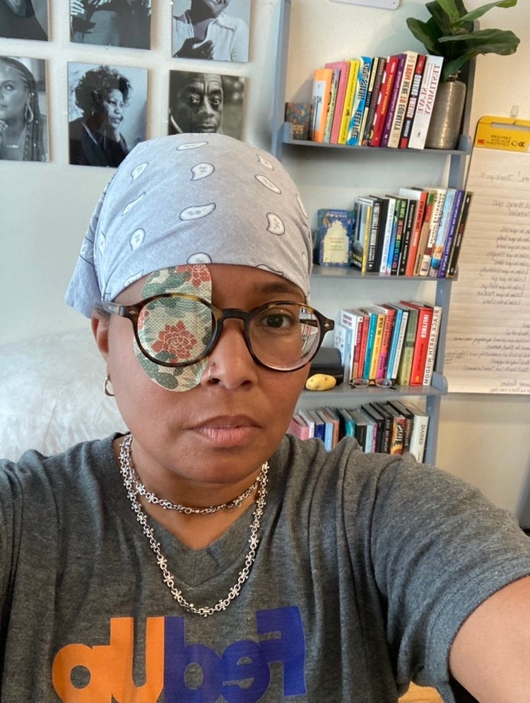 A picture of me wearing a grey t-shirt that says fed up, a grey scarf and a green patch with a cactus on it. In the background is my bookshelf and pictures of Black writers and other figures I admire. 