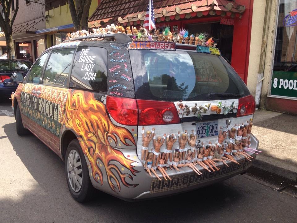 A minivan covered with Barbies, Kens and other toys, as well as odd paintings and mosaics. The window says TheBarbieLady.com