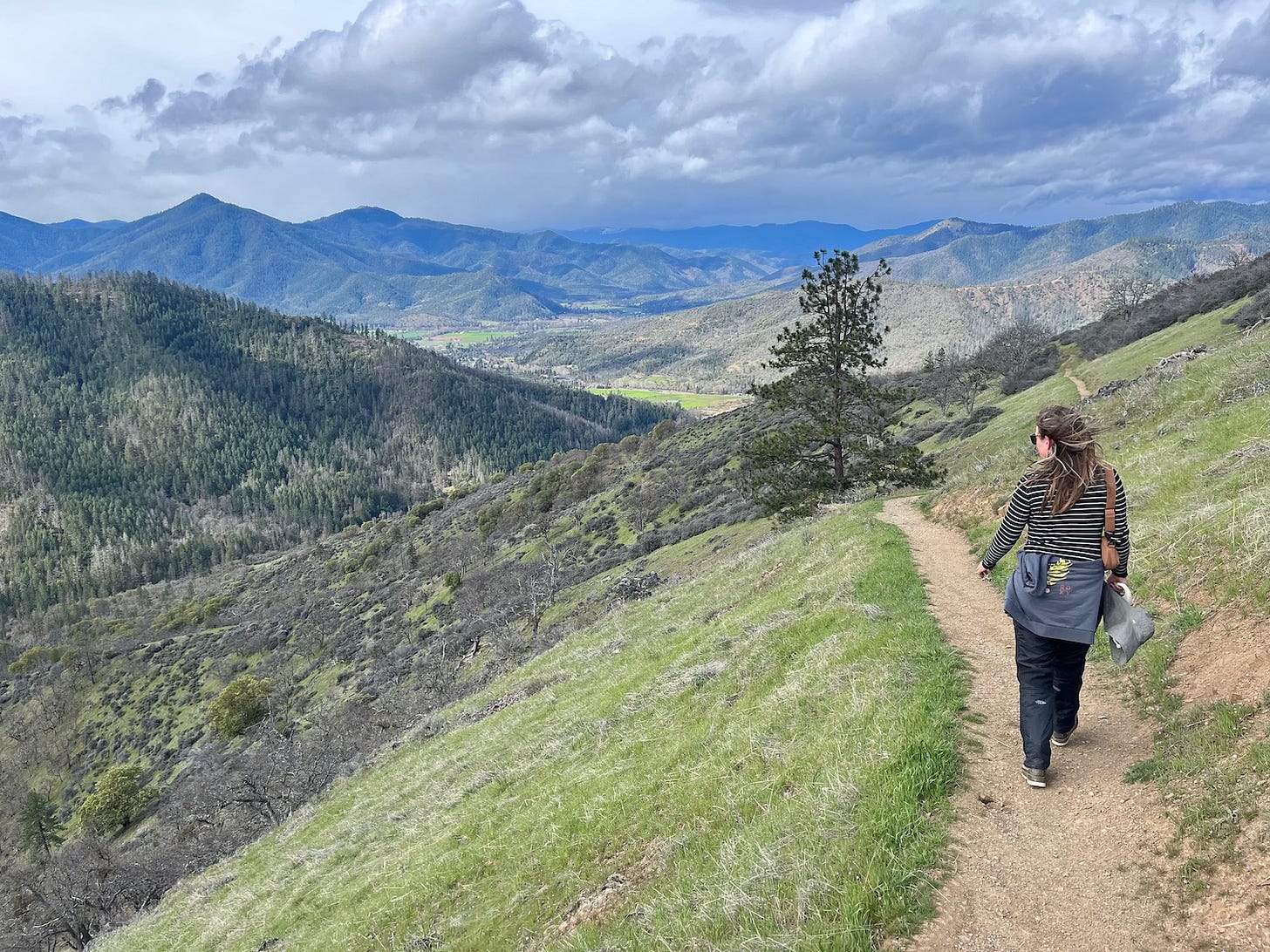 woman walks along a trail with steep grass on either side and mountains in the distance