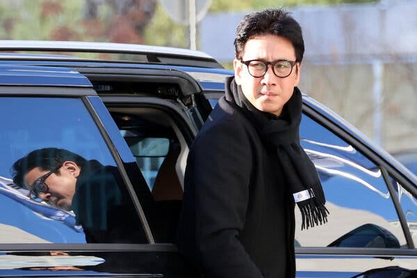 A man wearing glasses, and a black overcoat and black scarf steps out of a vehicle.
