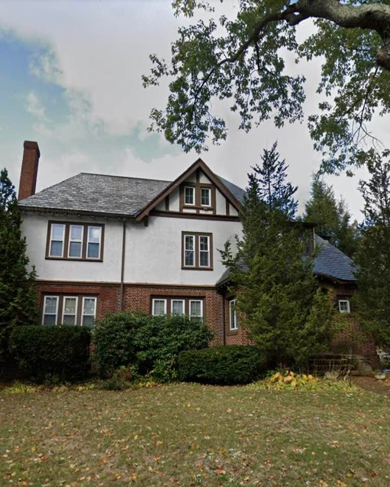 Photo of a three-story brick and stucco house with tall chimneys, a slate roof and many windows. It sits on a low rise; there is a small yard in front of it and shrubs and small trees on all sides. 