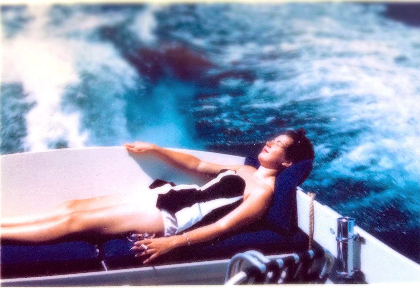 A young woman with dark hair wearing a black-and-white-striped, strapless bathing suit reclines on blue cushions in the back of a speedboat, white water and blue waves blurred in the background.