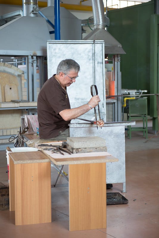 A man sits at a workbench and uses a metal tool to shape the molten glass at the end of a rod.