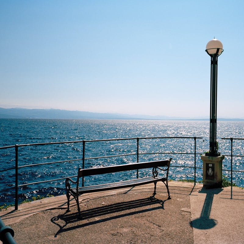 An empty bench beside a street light at the end of a peer overlooking a body of water with moutains in the distance.