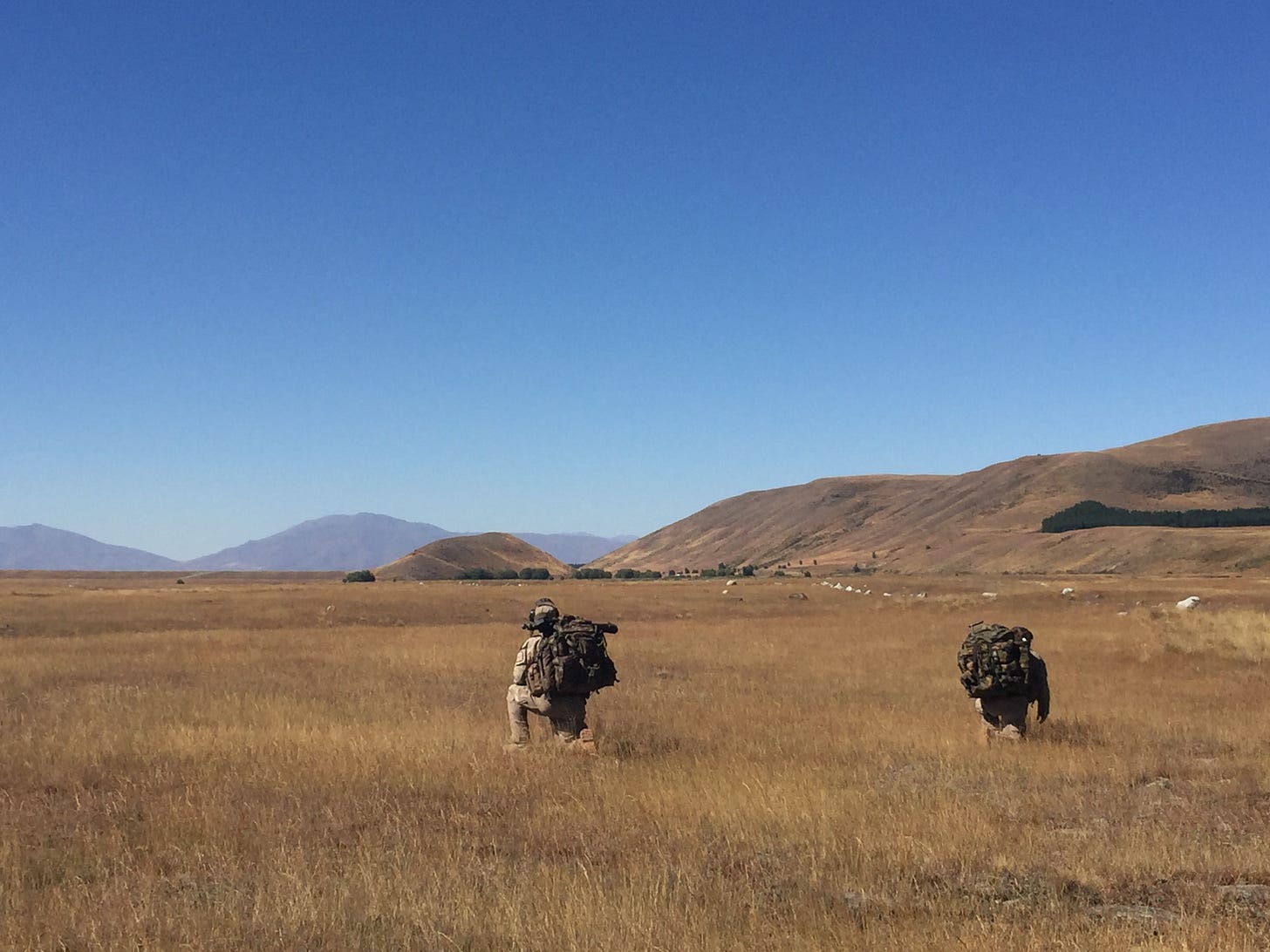 Brilliant blue sky fills the top half of the image, with the bottom half being brown tussock, and rolling hills in the mid-distance. Two NZ Army soldiers kneel in the foreground with their backs to the camera.