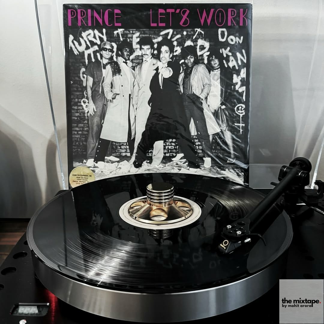 A vinyl record of Prince's single "Let's Work" is spinning on a high-end turntable. The record features a golden record weight in the center. The album cover, displayed in the background, shows Prince and his band against a graffiti-covered wall with vibrant pink lettering for the title. The setup suggests a dedicated vinyl collector's listening space.
