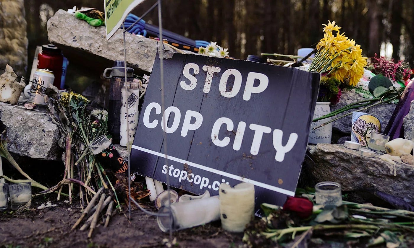 A photograph of a makeshift memorial altar in Weelaunee Forest, with many flowers, candles, and a sign that says STOP COP CITY.