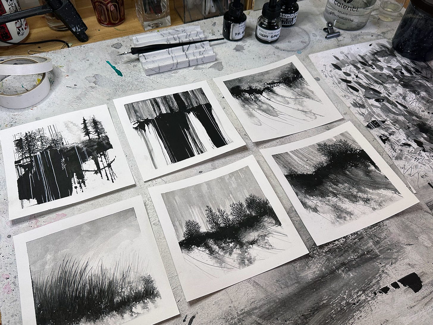 Photo of siz black and white ink paintings on my messy desk- the ppaintings are small, square, with white borders, and they are abstract landscapes in character- moody and atmospheric.