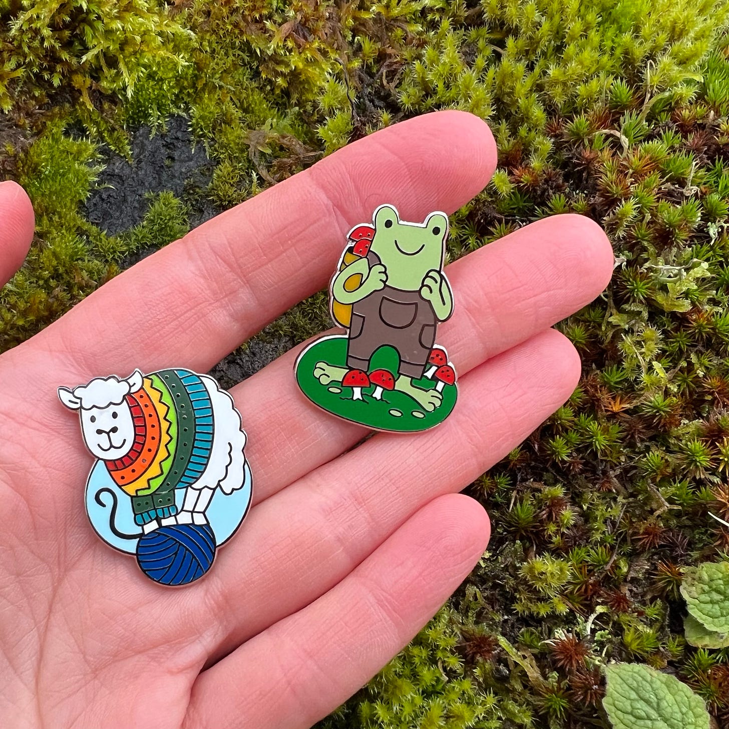Two enamel pins: a sheep in a rainbow jumper, and a foraging frog in dungarees