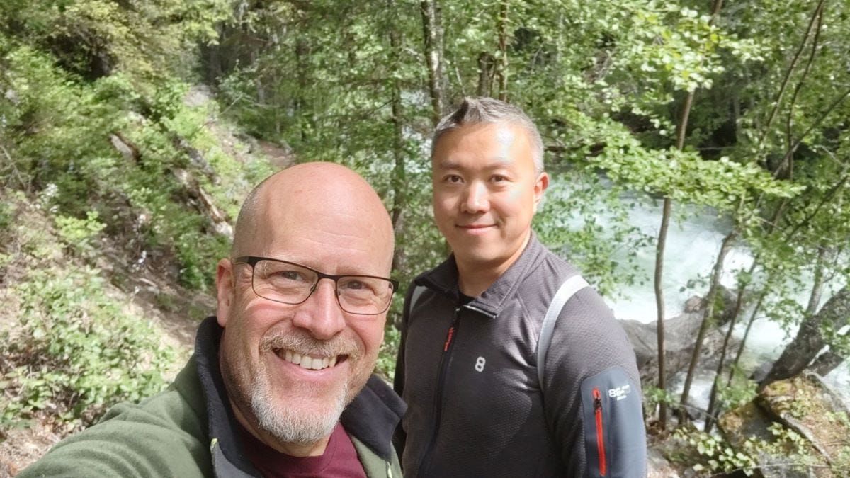 two men in a wooded natural environment with a rushing whitewater stream behind them