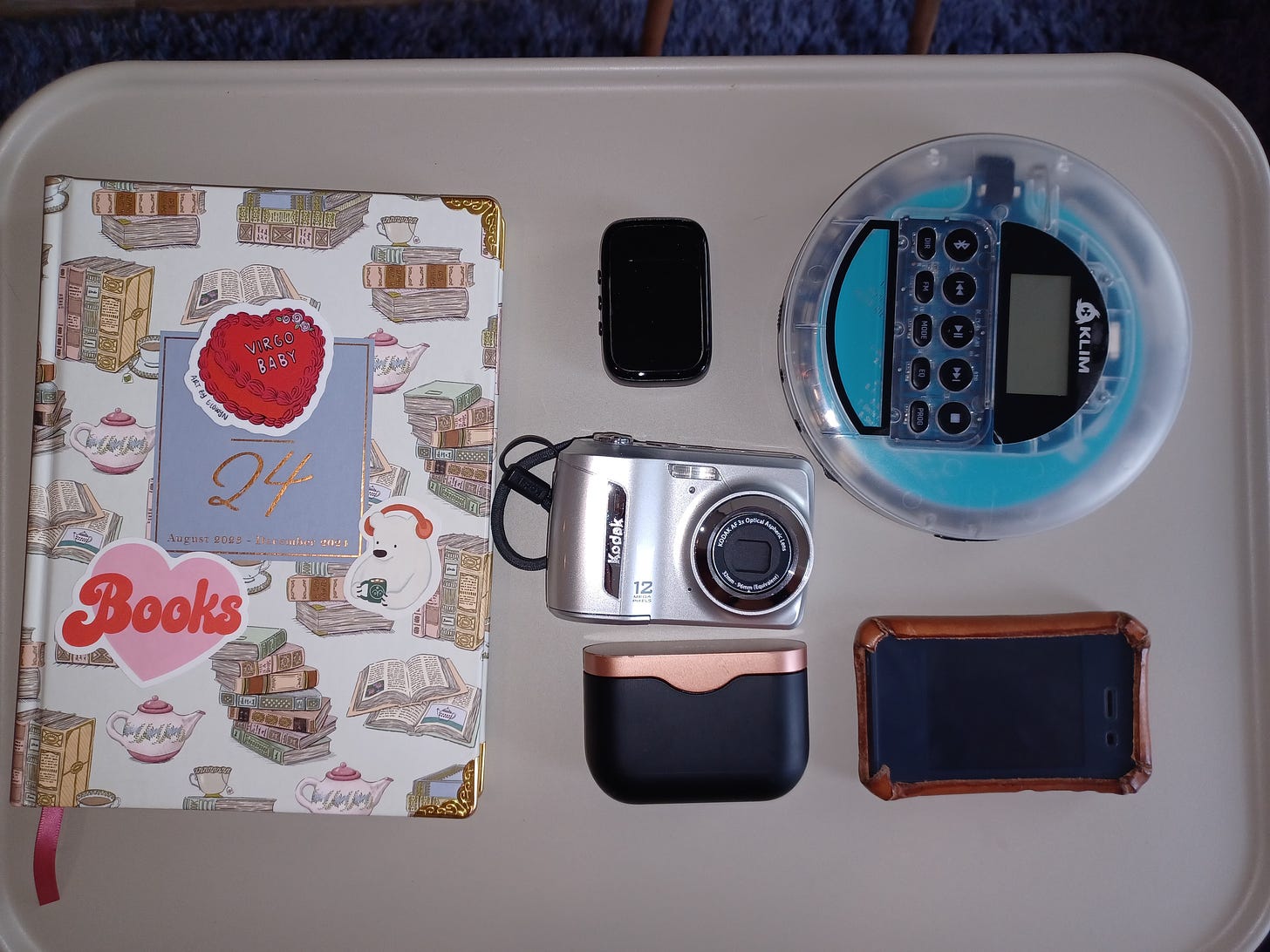 A picture of a hardbound paper planner, a digital Kodak Easyshot camera, bluetooth headphones, a Light Phone 2 in a case, a small mp3 player, and a Klim CD player.