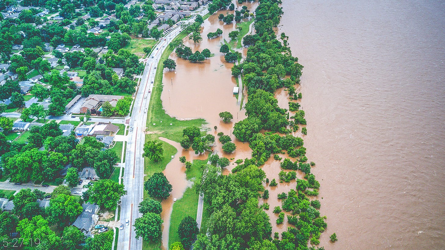 This aerial photo shows the tree-lined streets of Brookside meeting Riverside Drive, River Parks and the Arkansas River during the 2019 flood. The mostly flooded green space and swollen river dominate the center and right side of the image. Bright green clusters of treetops, which look like heads of broccoli, contrast boldly with the mud-orange floodwater. 