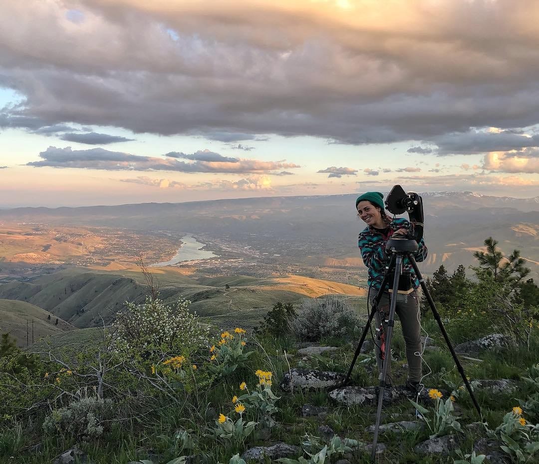 a scientist stands with a special camera designed to observe the night sky atop a mountain with valleys and a river behind her a scientist stands with a special camera designed to observe the night sky atop a mountain with valleys and a river behind her