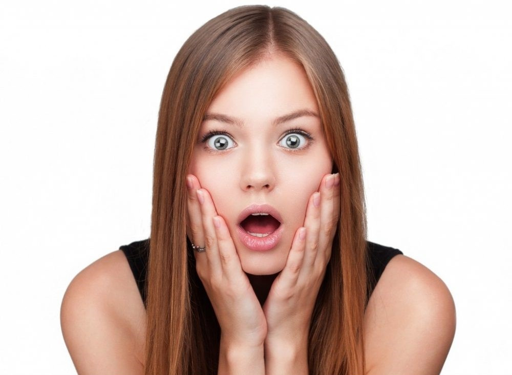 A young woman with long brown hair and wide blue eyes looks shocked, her hands pressed against her cheeks in surprise. A young woman with long brown hair and wide blue eyes looks shocked, her hands pressed against her cheeks in surprise.