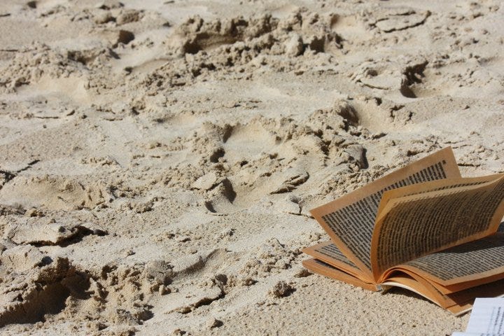 a close up of a sandy beach, and in the bottom right, a damp book, it's yellowed pages wet enough for dark lines of text to show through several pages. The paper curves, the pages looking like they're blowing in the wind.