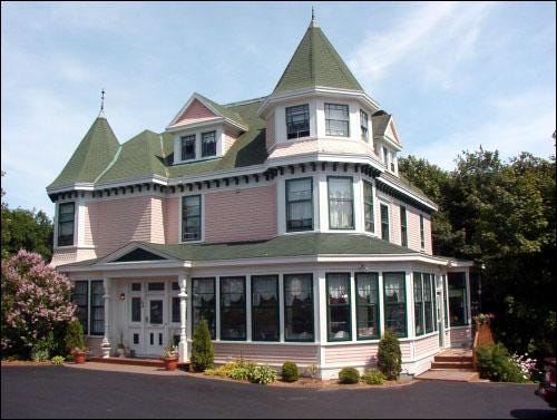 Photo of a Victorian-style old building with gables, turrets and a large wraparound porch, painted pink. 