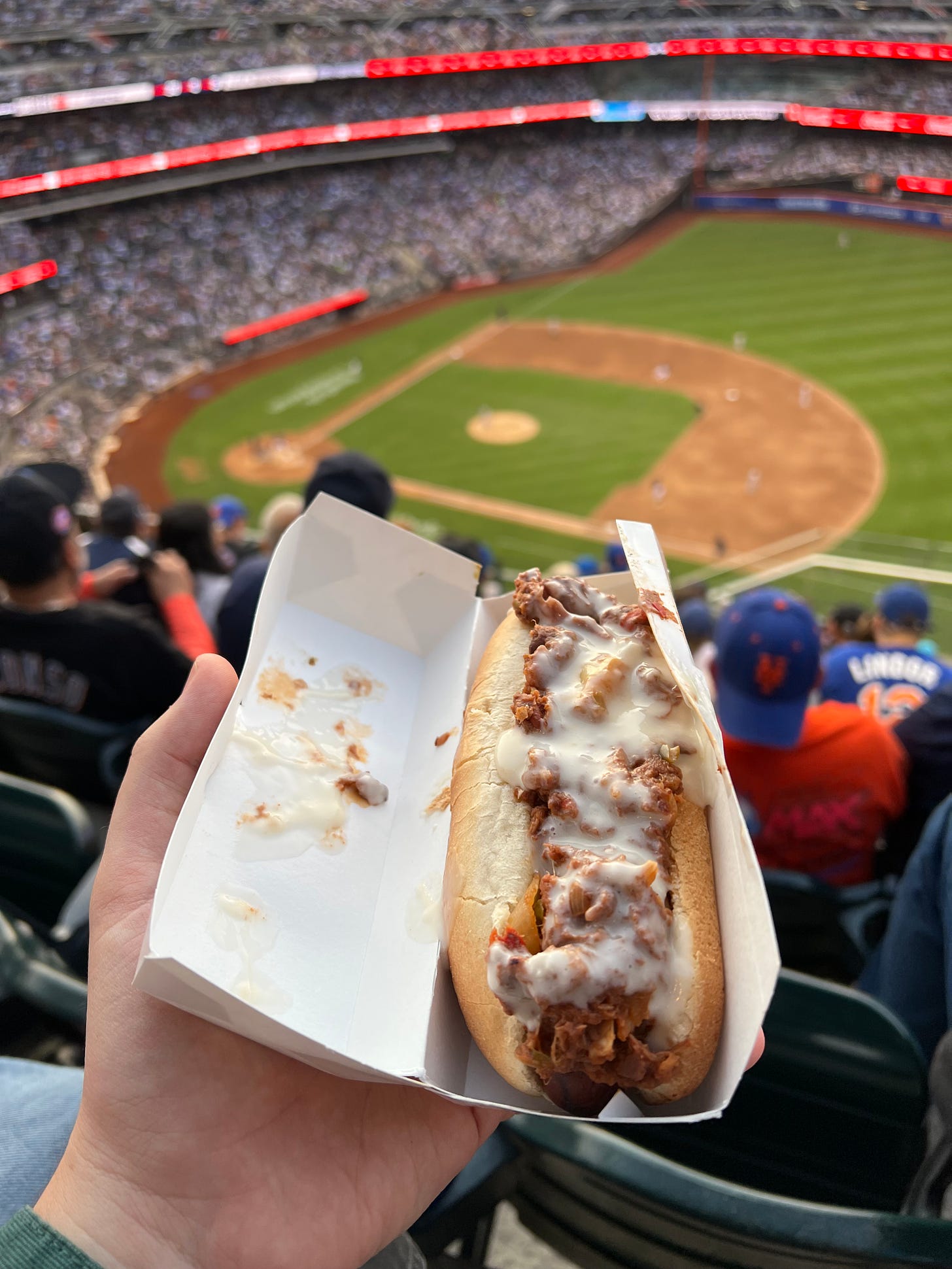 A Vegan City dog slathered in cheese sauce, blurred baseball game in background.