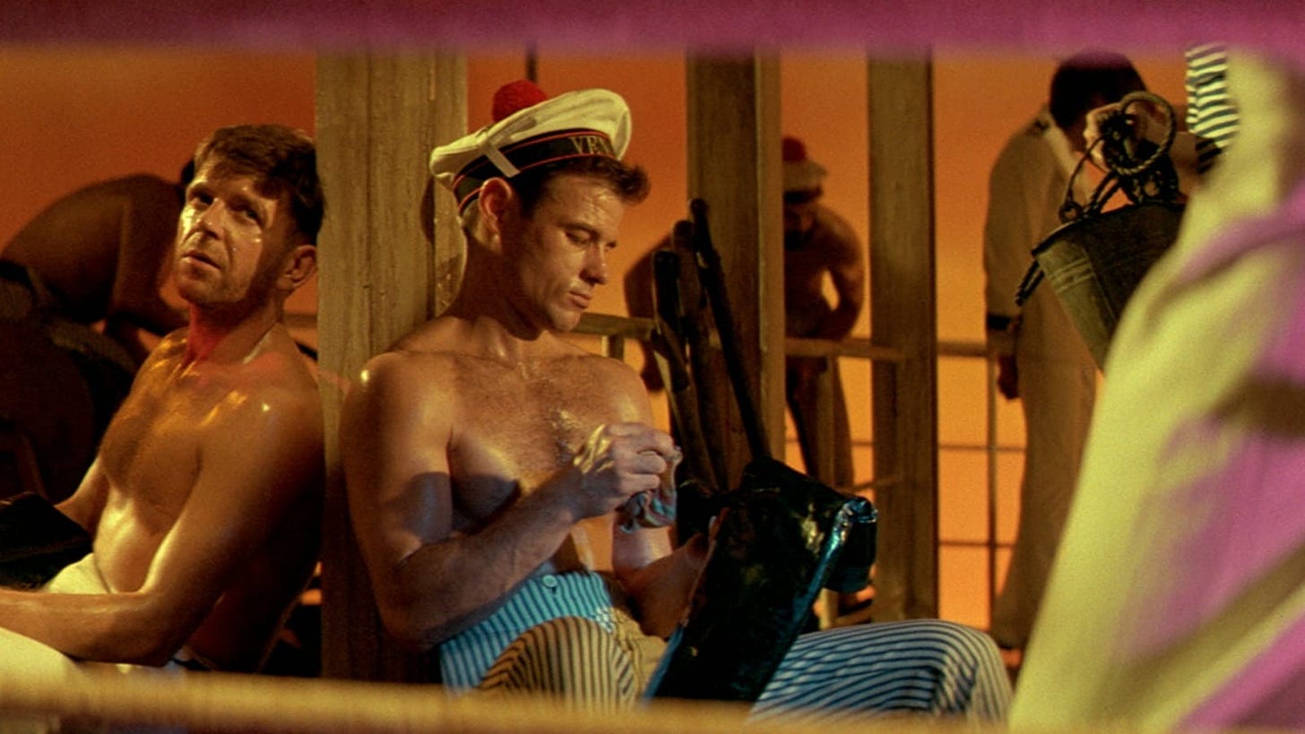 Movie still from Querelle. A topless sailor sits on deck next to other topless men.