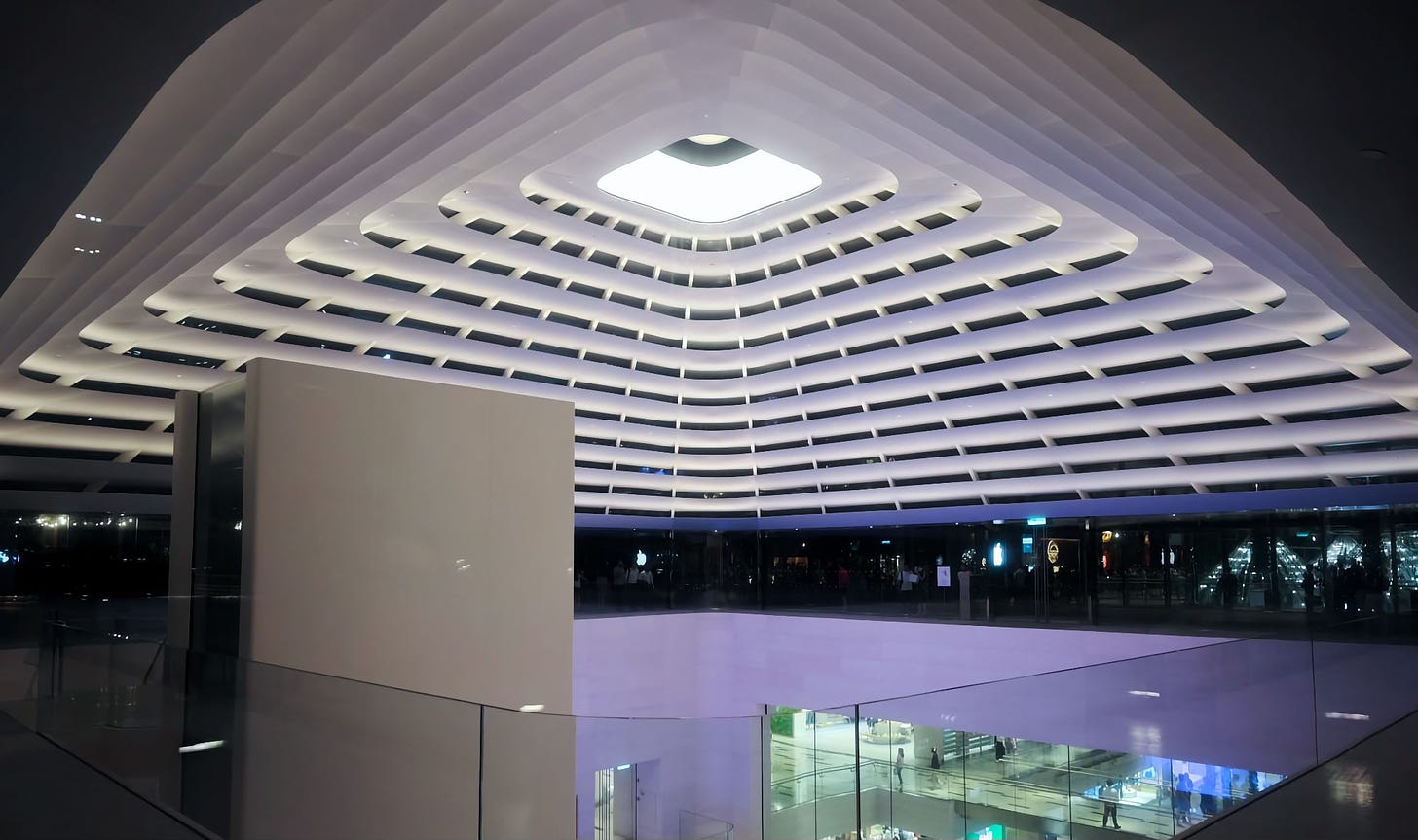The interior of the canopy at Apple The Exchange TRX at night. At the roof oculus, a lighting element illuminates the store below.