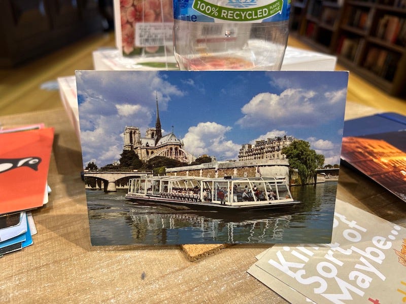 A postcard propped up against a water bottle. It shows a picture of a tour boat passing Notre-Dame.