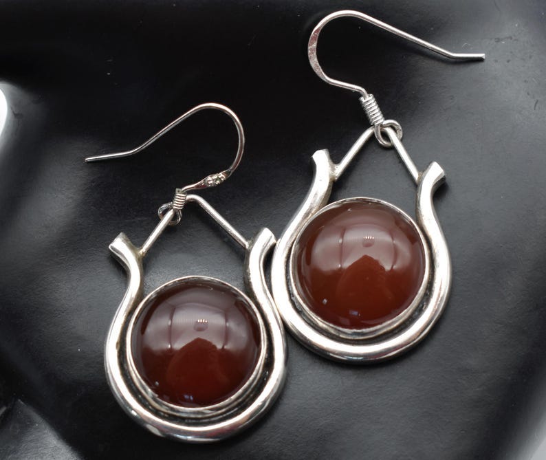 May include: A pair of silver dangle earrings with brown cabochon stones. The earrings have a teardrop shape with a curved design.