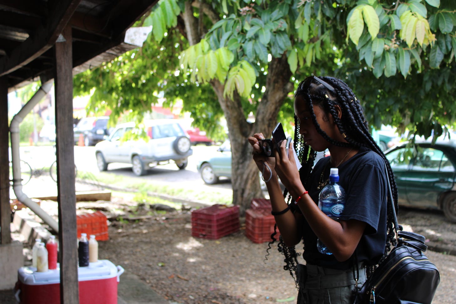 The image depicts a young woman pointing a photo camera at something out of frame. In the background stand a tree and a row of parked cars. To the left a wooden pillar supports a porch roof.