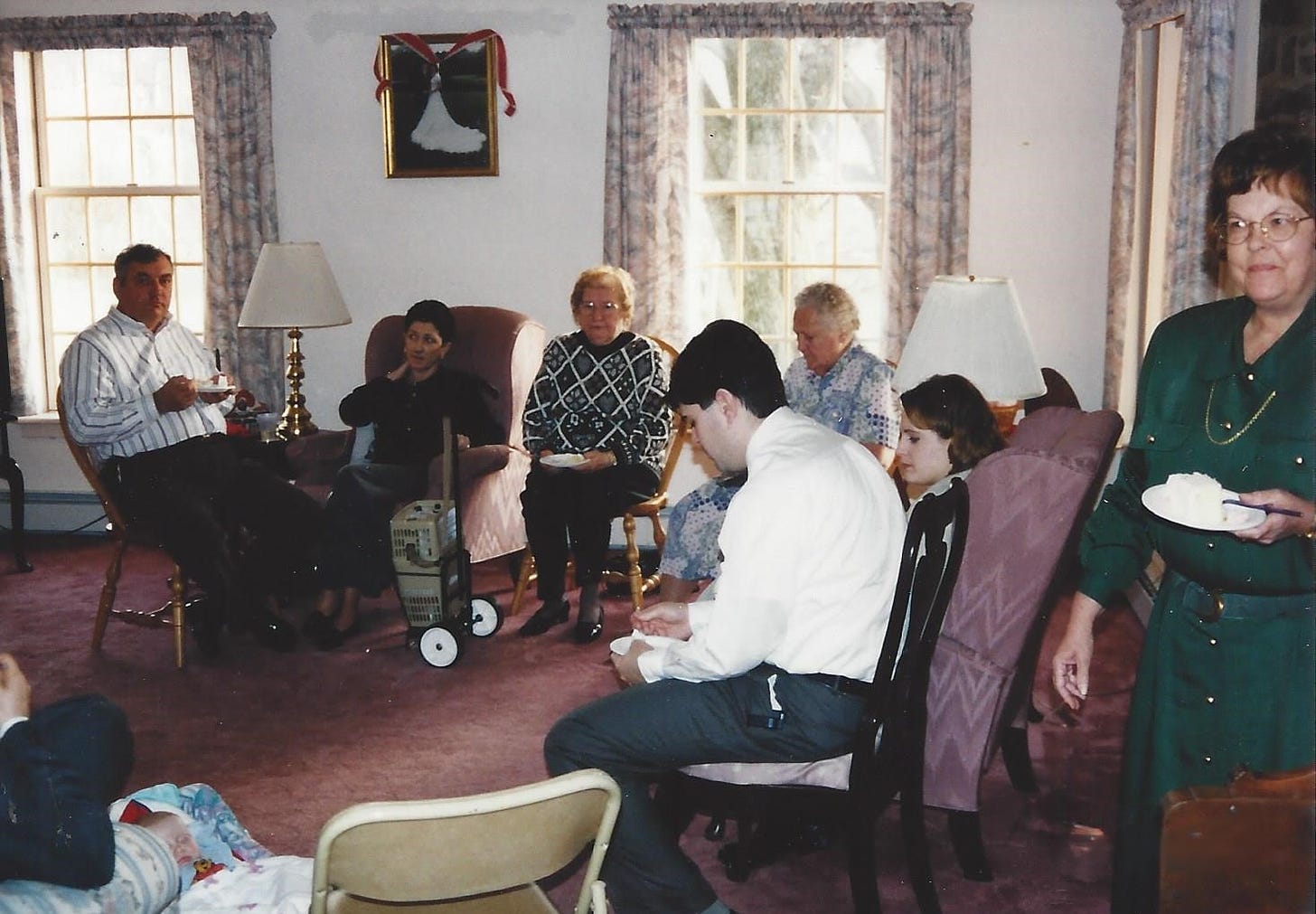 Numerous relatives are gathered in a living room facing an empty folding chair