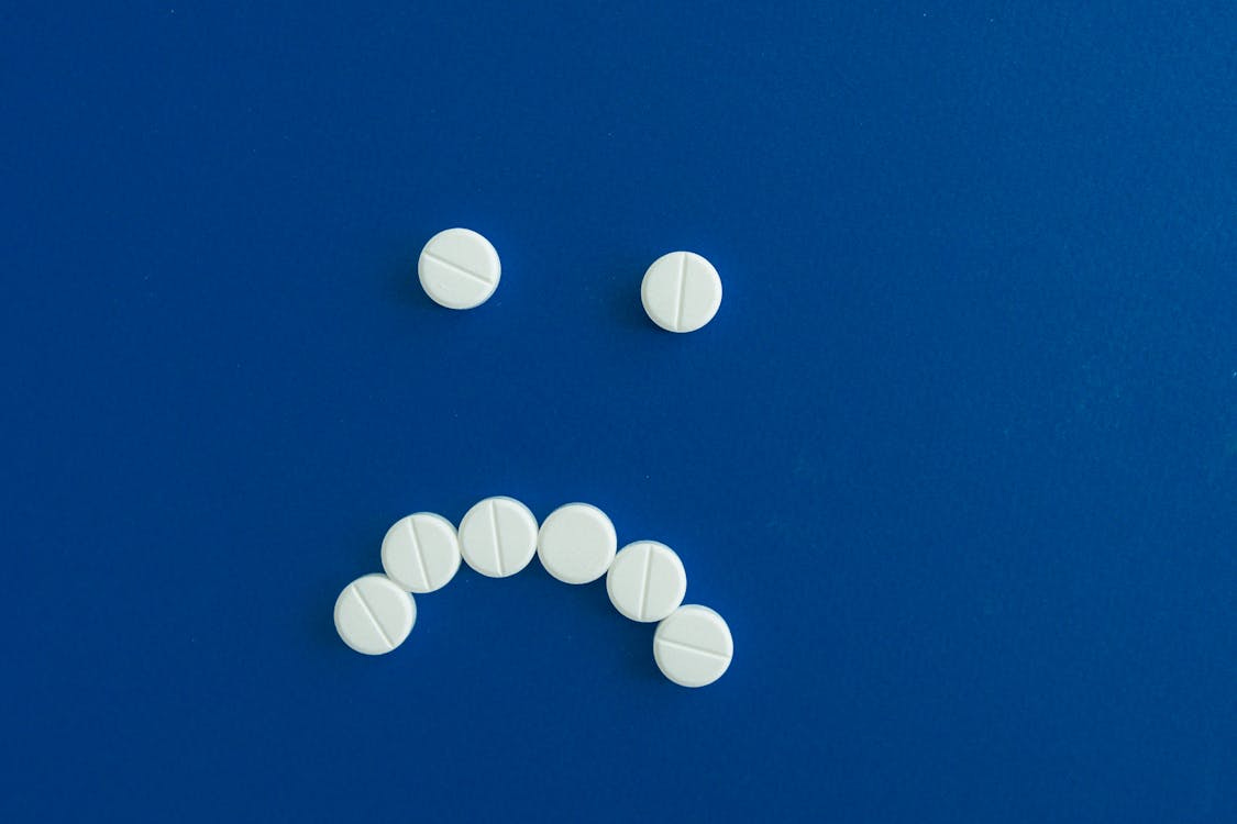 Free White pills arranged in a sad face shape on a solid blue background. Stock Photo