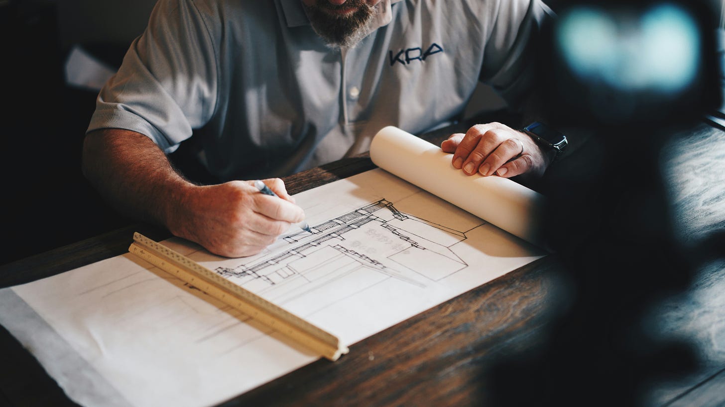 A bearded person draws a house plan with a paper and pencil.