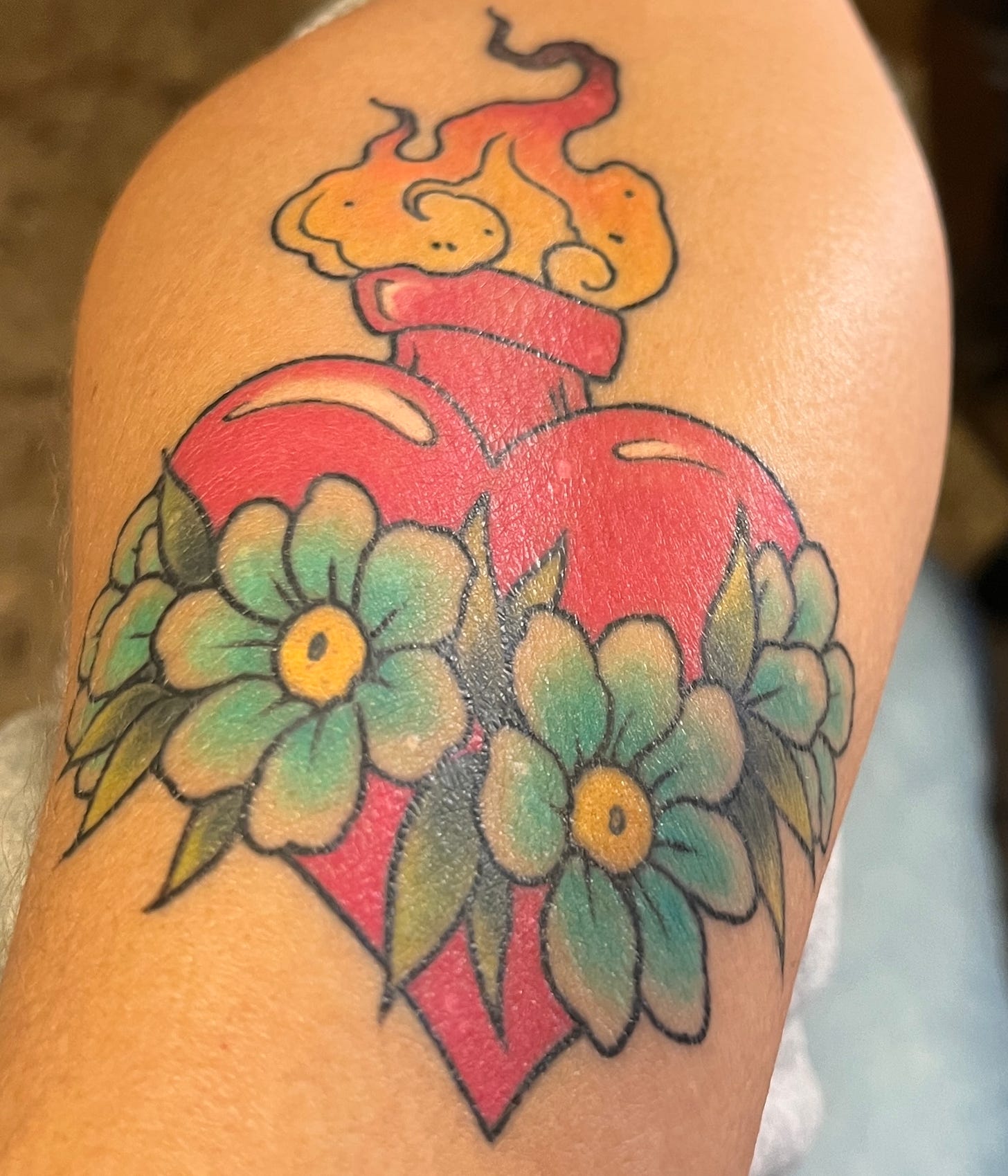 A tattoo on a tan-colored forearm of a red heart crossed with florals and with flame coming out of the top.
