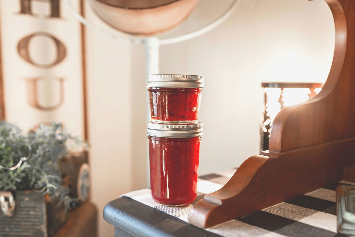 a larger and smaller ball-style jar on top of eachother filled with a red jam or jelly on the edge of a black countertop, country-style decorations in background