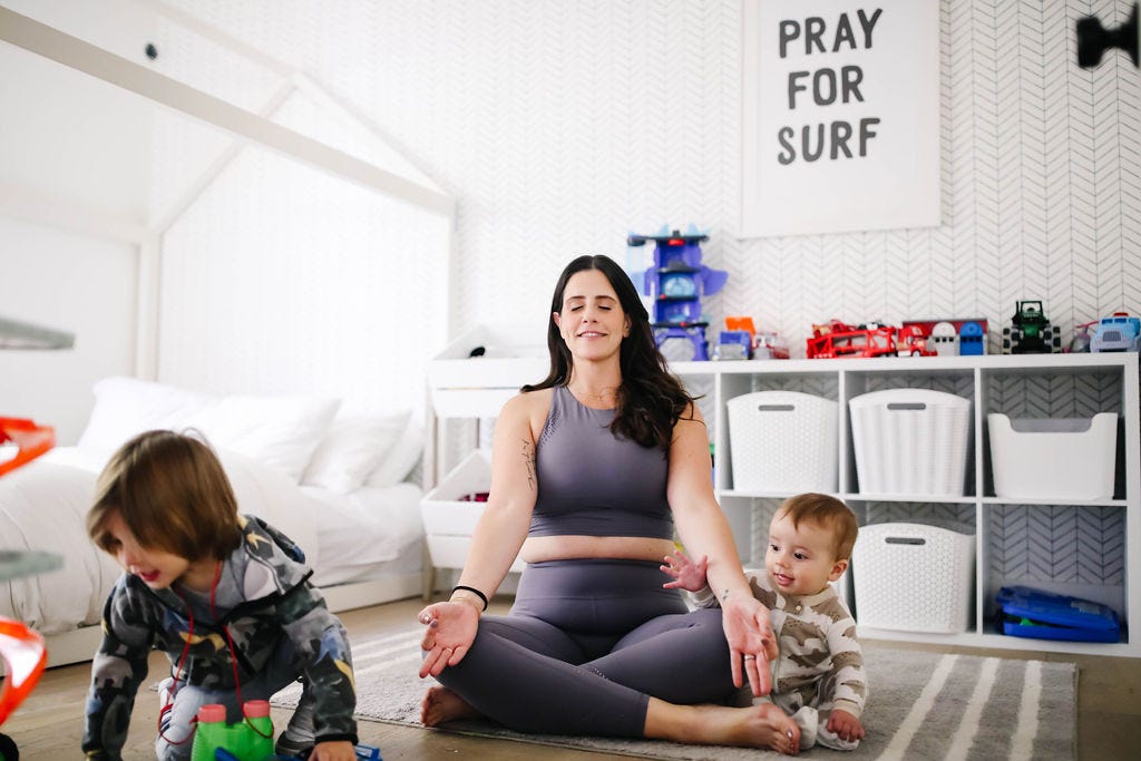 Sarah Ezrin seated in meditation in a child's bedroom with a toddler and a baby playing around her