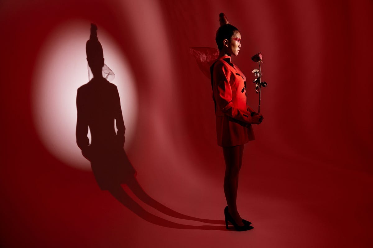 Woman in a red dress holding a red rose illuminated from the front, shadow on the wall behind her