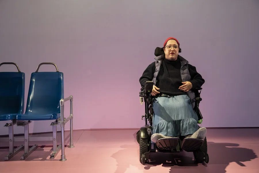 A color performance image of a femme using an electric wheelchair. She wears comfortable-looking slippers, a denim skirt, a black jacket, a vest, a red hat, and glasses. The background is a light purple, and she is by two metallic chairs.