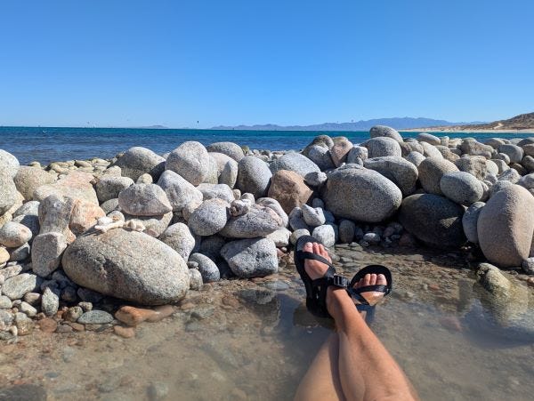 my legs, crossed at the ankle and wearing strappy black sandals in a shallow pool surrounded by rocks, with the deep blue sea beyond