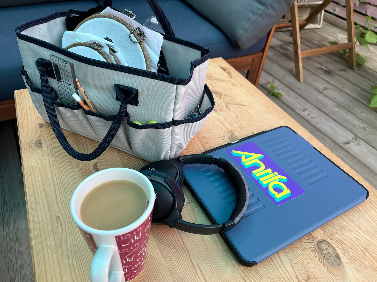  Photo of a rectangular wooden table on a grey wooden deck. A light grey craft bag sits on the table, several embroidery hoops inside with art supplies poking out of pockets all aroudn the outside of the bag. A cup of tea in a magenta and white mug sit on the table next to some headphones and a tablet with a vibrant Antifa sticker on it.