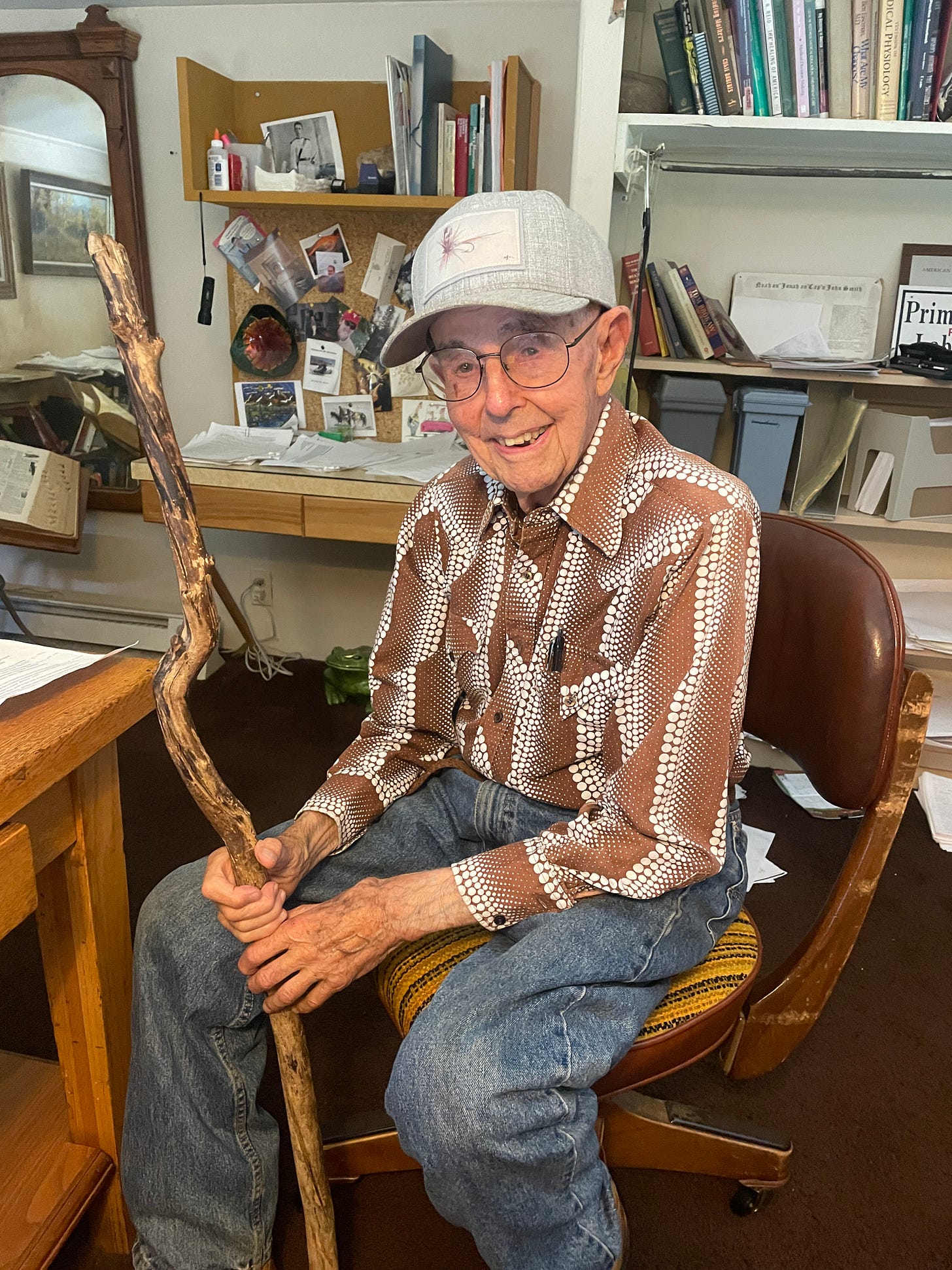 My Dad, Poppa Stu at age 99. He is in a classic 70s brown polkadotted shirt, a fly-fishing hat, and holding a gnarled old walking stick. He is a white man, smiling at the camera, sitting on a brown chair at an office desk with books behind him.