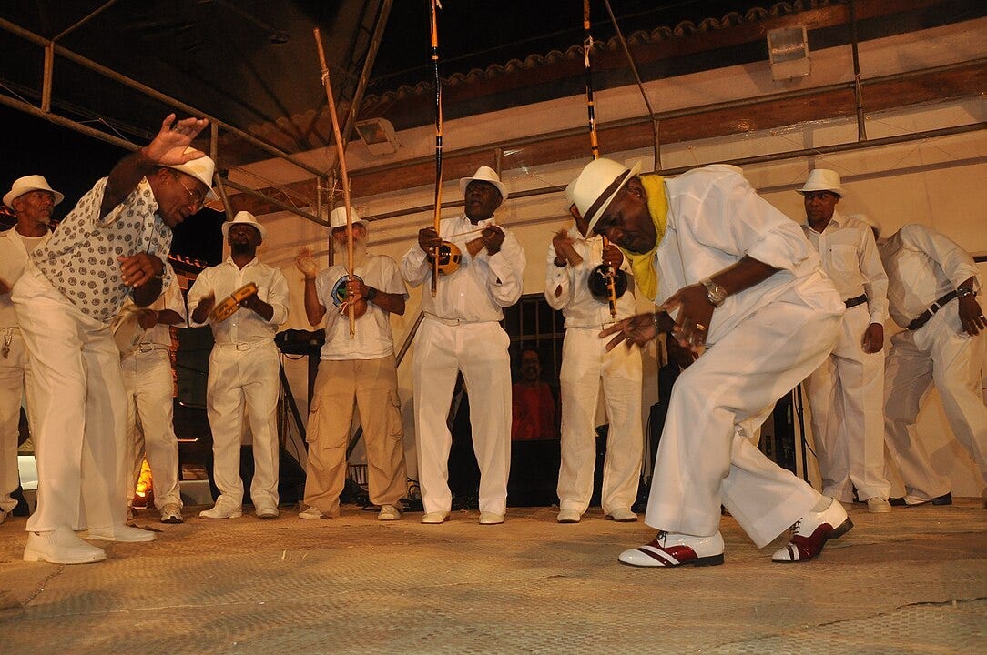 A Capoeira Angola player calls a “chamada” by raising his hand. His partner in the game begins his approach. Note the immaculate white clothing. 