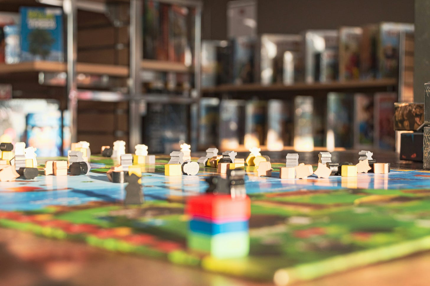 focused shot of a board game in action. wooden pieces sit around the colorful board with green, red, and blue areas. the background is blurry with other boxes and games behind the table.