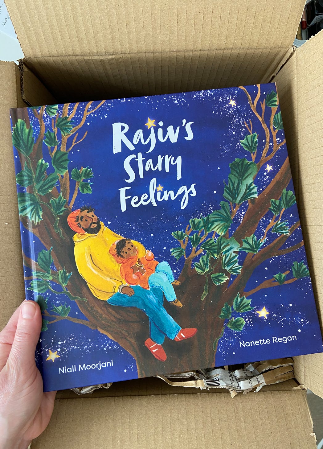 hand holding a copy of Rajiv's Starry Feelings by Niall Moorjani, illustrated by Nanette Regan. The cover shows a father and son hugging in the branches of a tree surrounded by stars in the night sky