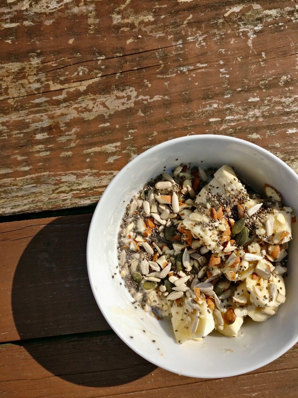 a bowl of oatmeal with bananas, nuts, and seeds on a wooden table in the sun