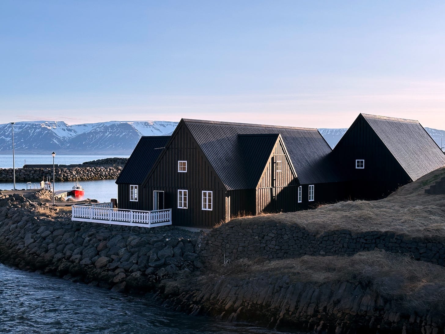 Evening sunlight in Hofsos, Iceland.  There are several black painted buildings with our rental apartment on the left side.  A red boat is in the harbor, a river runs across the foreground into the fjord and snow capped mountains are across the fjord.  The sky is just begining to turn pink.