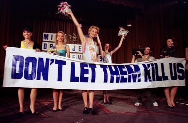 Image shows a picture of a beauty pageant winner ceremony. The winner, Irena Nogić, is holding a bouquet of flowers above her head while the other contestants stand beside her. The are holding a sign that says "don't let them kill us."