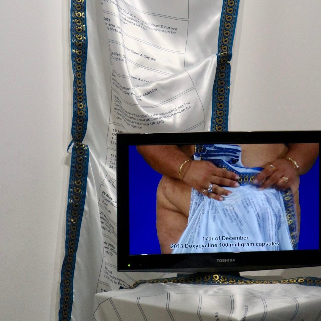 A still from the installation of a film work on a monitor in front of a piece of fabric. Set against a stark blue background, the camera focuses on the bare hands and lower torso of the artist, an unclothed woman with a pale brown skin tone and full figure, who wears gold bangles and rings. The artist holds before her intimate parts, which remain covered, a long, almost never-ending piece of fabric printed with a list of medical prescriptions, including dates and medications. The silk-like fabric has an intricate and detailed border in blues and golds, with a repeating swirling pattern that stretches its full length. The print is in black text on a white background. Throughout the film the artist concertinas the printed list in their grip, making fold on fold that hangs before them, gradually gathering up into bunched folds that seem difficult to manage. A slow yet obsessive process that is repeated until two thirds of the way through, when the artist starts to reverse the process, gradually releasing the folds. From this point on, the artist alternates between steadily gathering up the folds and releasing them in a process of painstaking repetition.