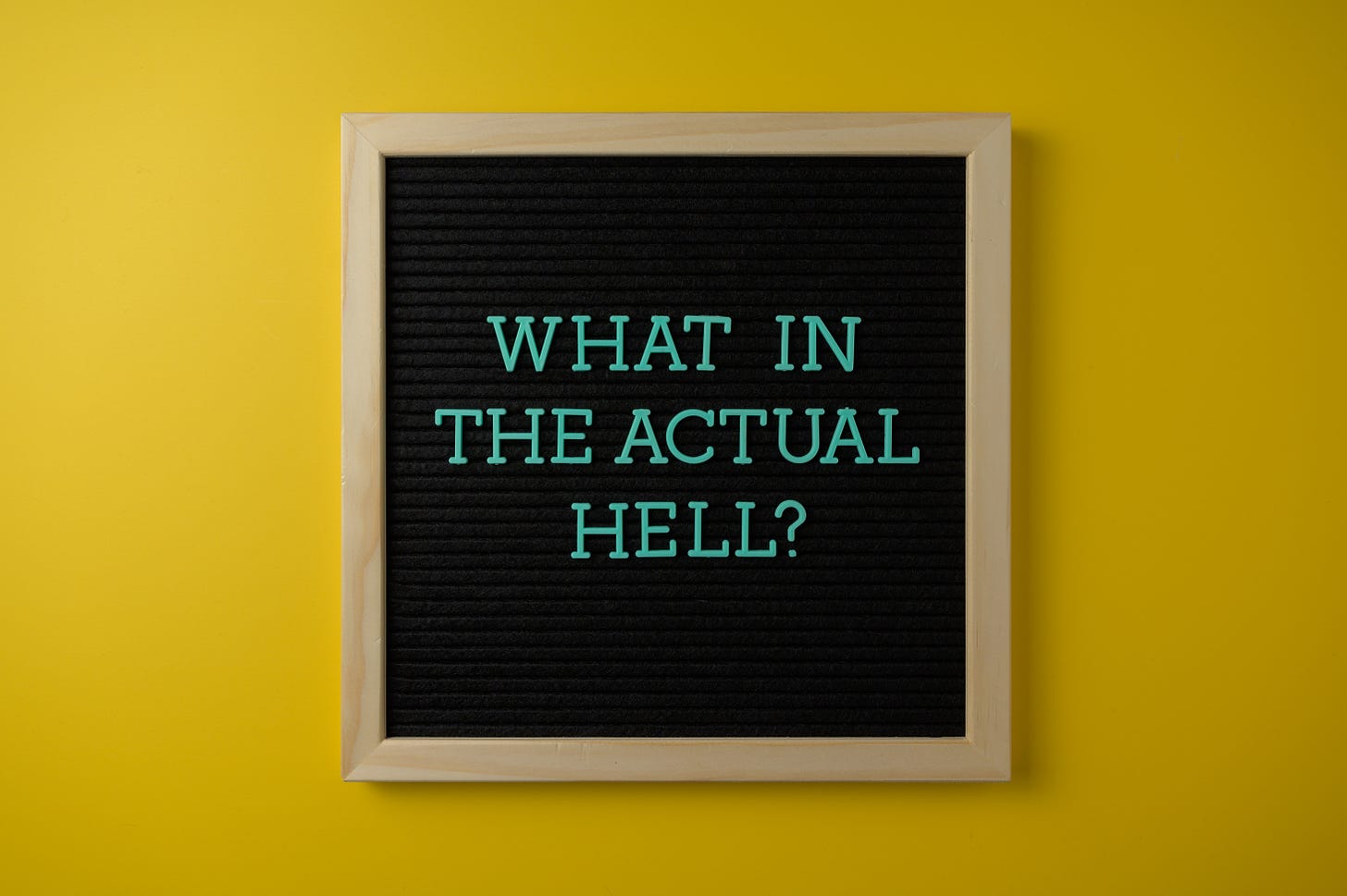 a blackboard with the words what in the actual hell written in teal - the board sits on a yellow wall