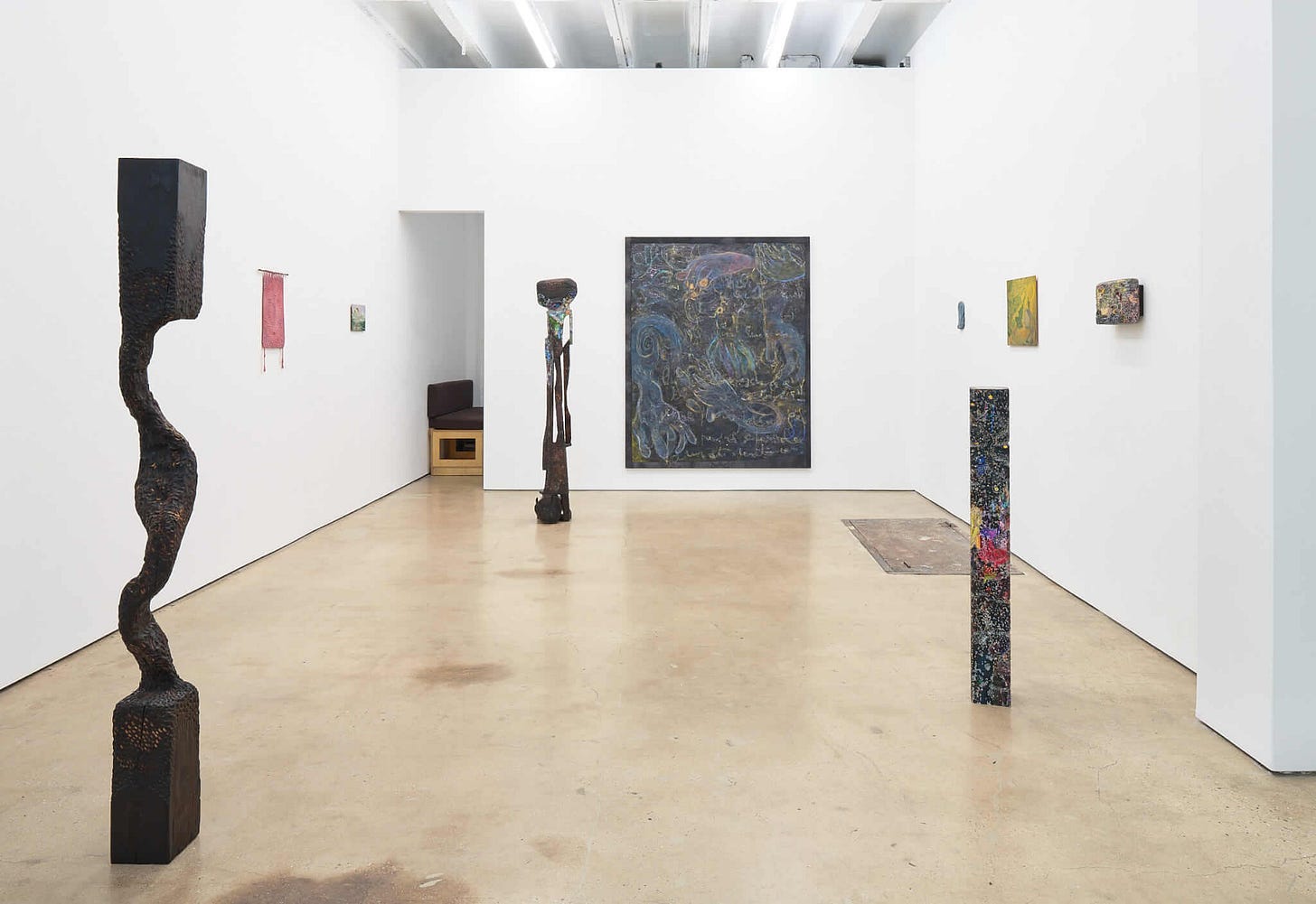 View of the gallery: white walls, three freestanding sculptures by Borba, that are made out of dark materials like wood but with some abstract paint on them; small, book-size paintings and fabric works line the walls, along with one large painting in the back that shows swirling hands in a black, purple, and blue palette. View of the gallery: white walls, three freestanding sculptures by Borba, that are made out of dark materials like wood but with some abstract paint on them; small, book-size paintings and fabric works line the walls, along with one large painting in the back that shows swirling hands in a black, purple, and blue palette.